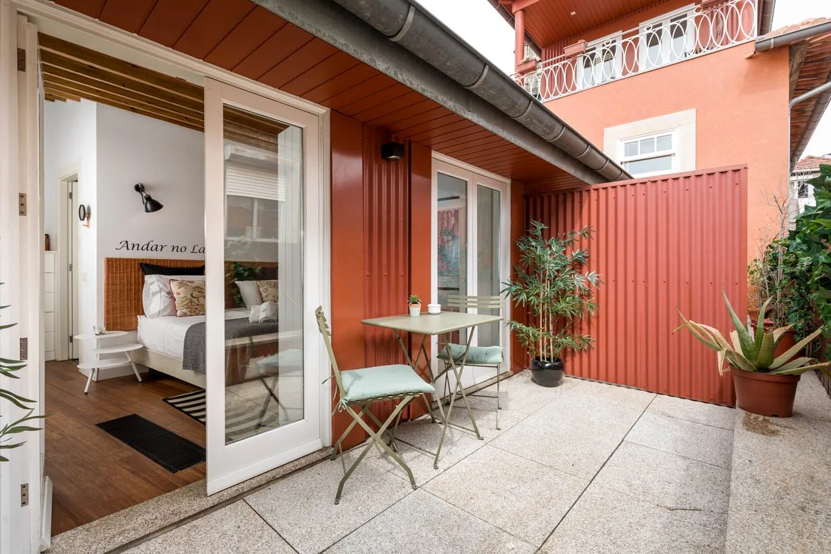 Varanda com mesa e duas cadeiras, vaso com plantas, parede de metal ripado, porta de vidro com vista para quarto decorado, muro ao fundo.