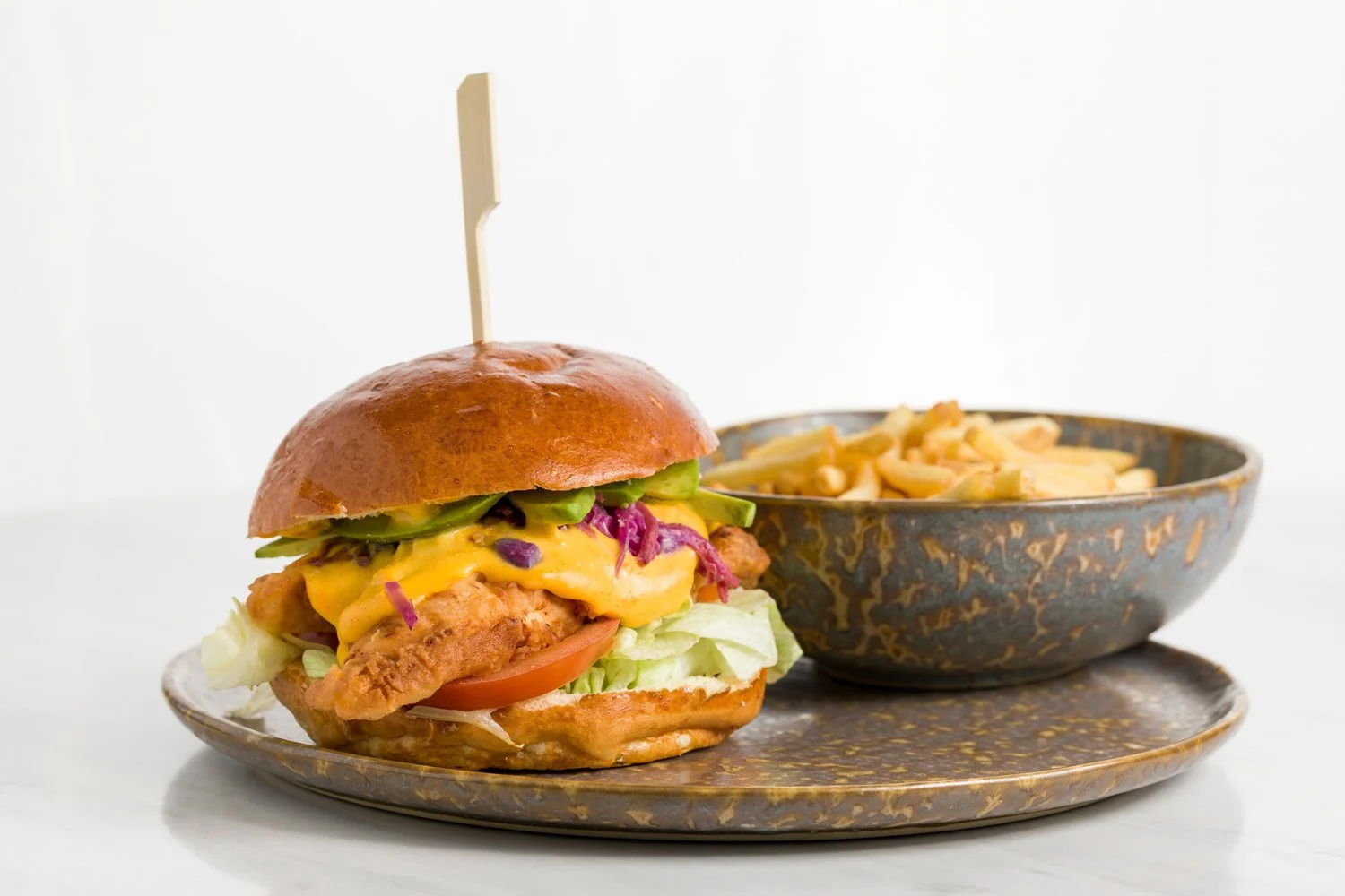 Sanduíche de frango crocante com queijo, alface, tomate, abacate, repolho roxo em pão brioche, acompanhado de uma tigela de macarrão gratinado, tudo em um prato de cerâmica marrom decorativa.