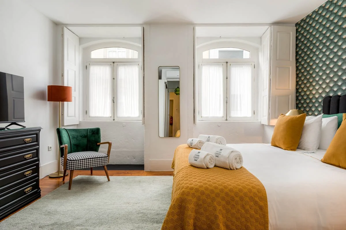 Quarto com duas janelas, cama com roupa branca, travesseiros amarelos, um cobertor amarelo, toalhas brancas na cama, espelho pendurado na parede, poltrona verde com estofado xadrez preto e branco, cômoda preta com TV, abajur laranja e parede decorada com papel de parede de padrão geométrico.