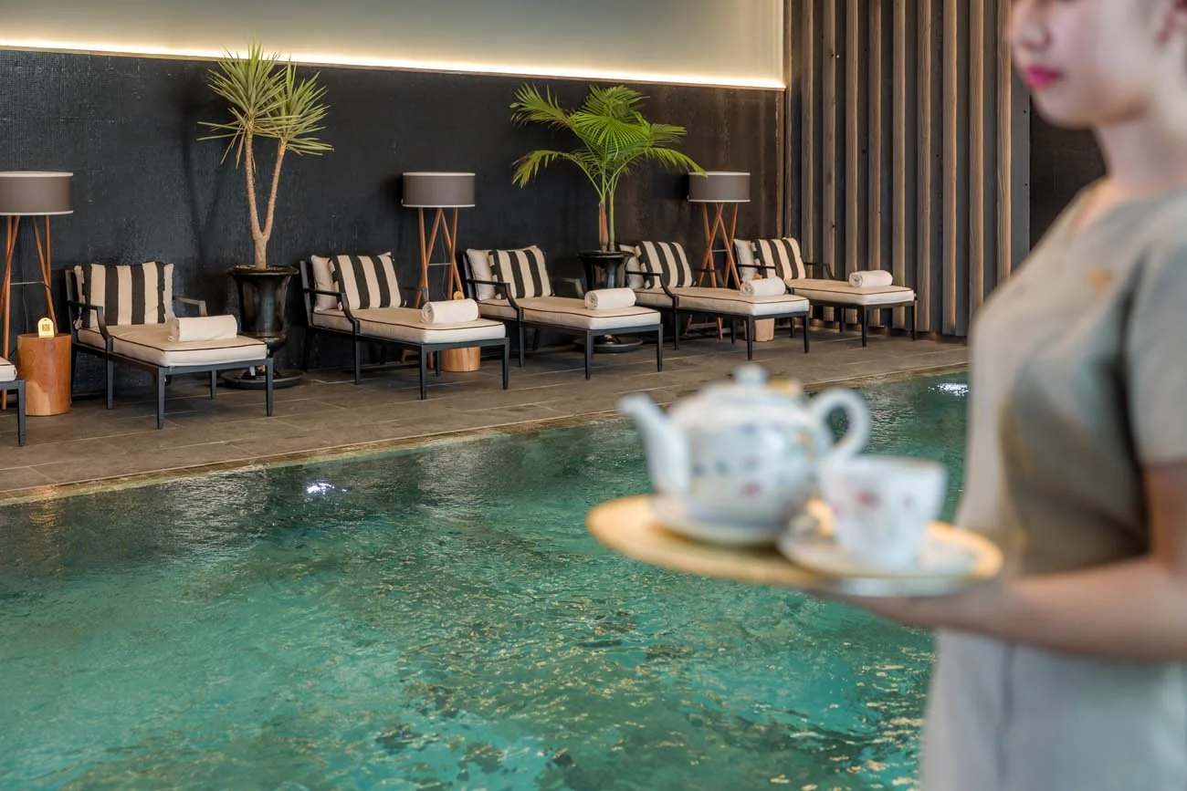 Pessoa segurando uma bandeja com bule e xícaras ao lado de uma piscina interior, com cadeiras de descanso e plantas ao fundo.