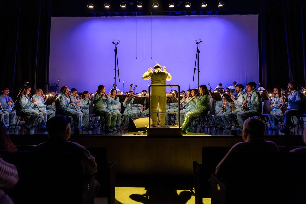 Orquestra de músicos tocando em palco com um maestro conduzindo, iluminação azulada, espectadores assistindo ao espetáculo.