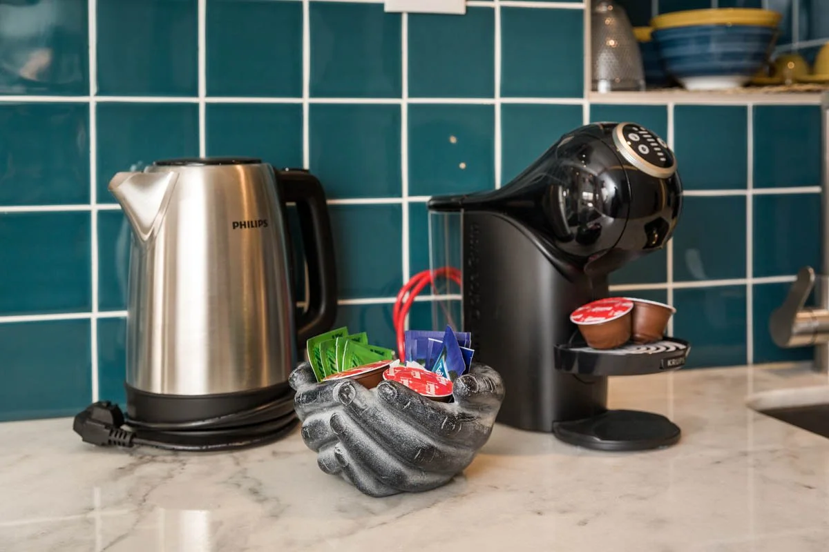 Máquina de café de cápsulas, chaleira elétrica, e um recipiente com cápsulas de energéticos em uma bancada de cozinha com revestimento de azulejos azul.