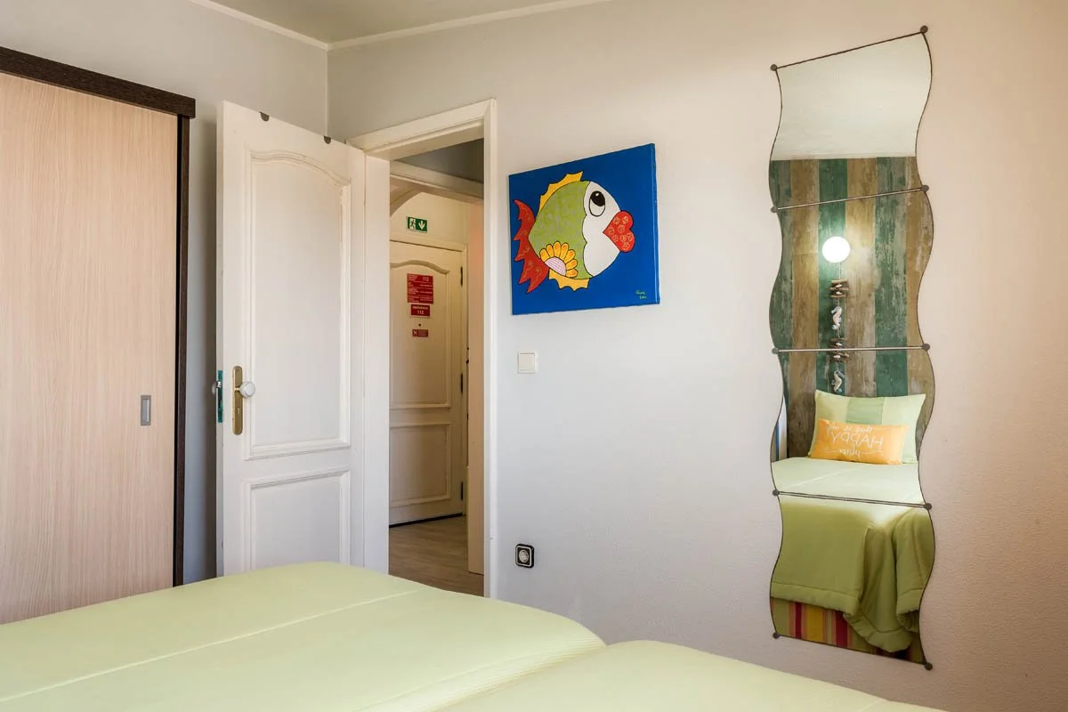 Quarto com cama de lençol amarelo, espelho ondulado na parede, uma pintura de peixe no lado direito, e uma porta de madeira branca aberta.