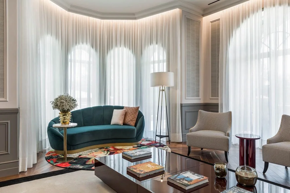 Sala de estar com janelas grandes com cortinas brancas, um sofá verde escuro, duas poltronas cinza, uma mesa de centro preta com livros, uma luminária de pé e uma mesa lateral com vaso de flores.