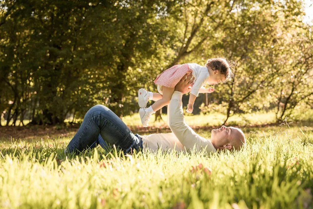 Homem deitado no gramado brincando com uma menina no parque ao ar livre, cercados por árvores verdes.