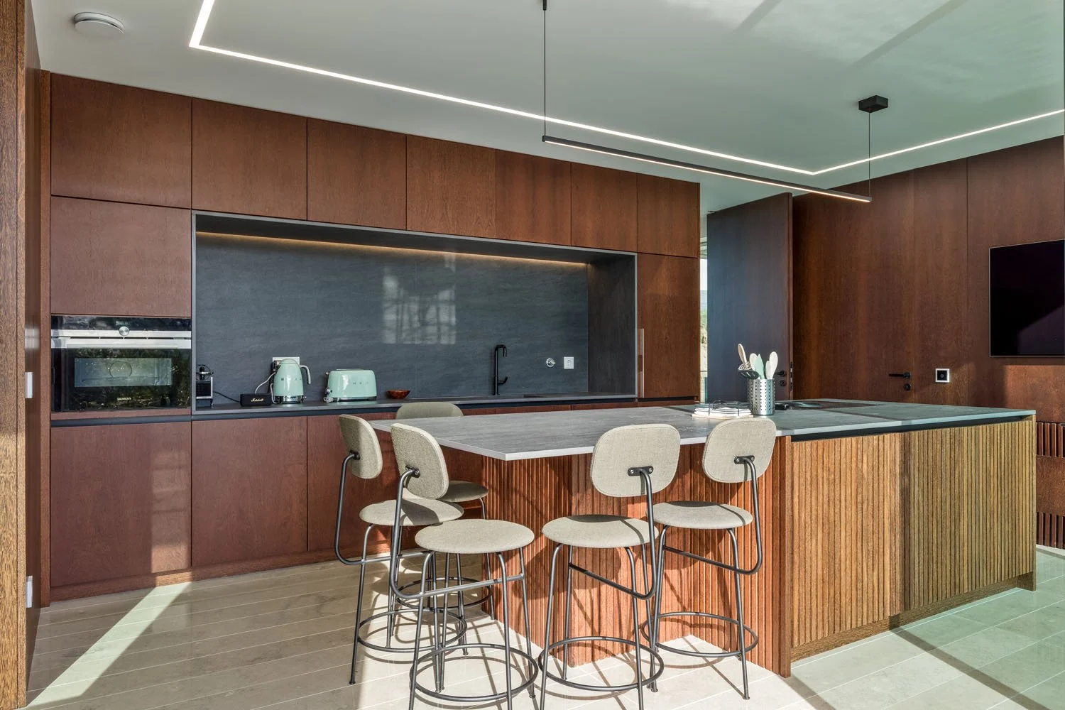 Cozinha moderna com armários de madeira, ilha com bancada cinza, quatro banquetas com assentos confortáveis, utensílios de cozinha em cores suaves, como liquidificador e torradeira, parede de fundo em azul escuro, iluminação embutida no teto e televi