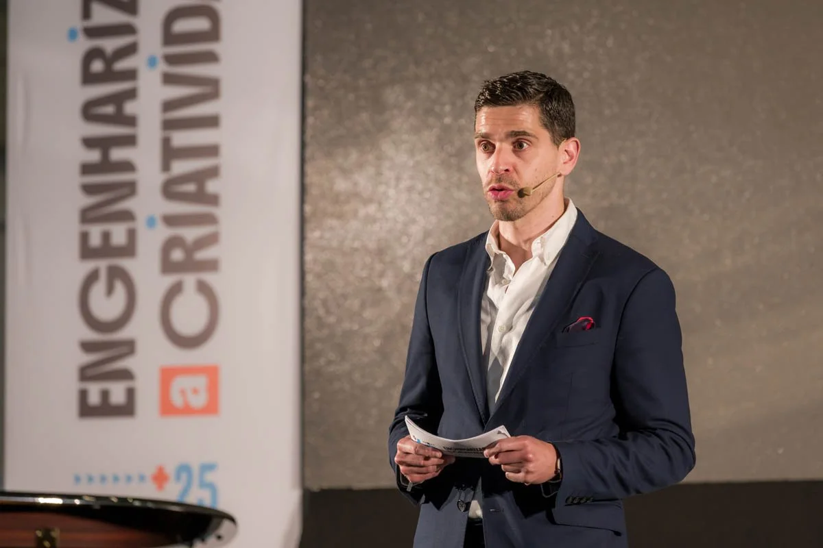 Homem falando em uma palestra com um microfone na orelha, vestindo terno azul escuro e segurando papéis, com uma placa ao fundo com o texto 'ENFRENTANDO O CRIME'.
