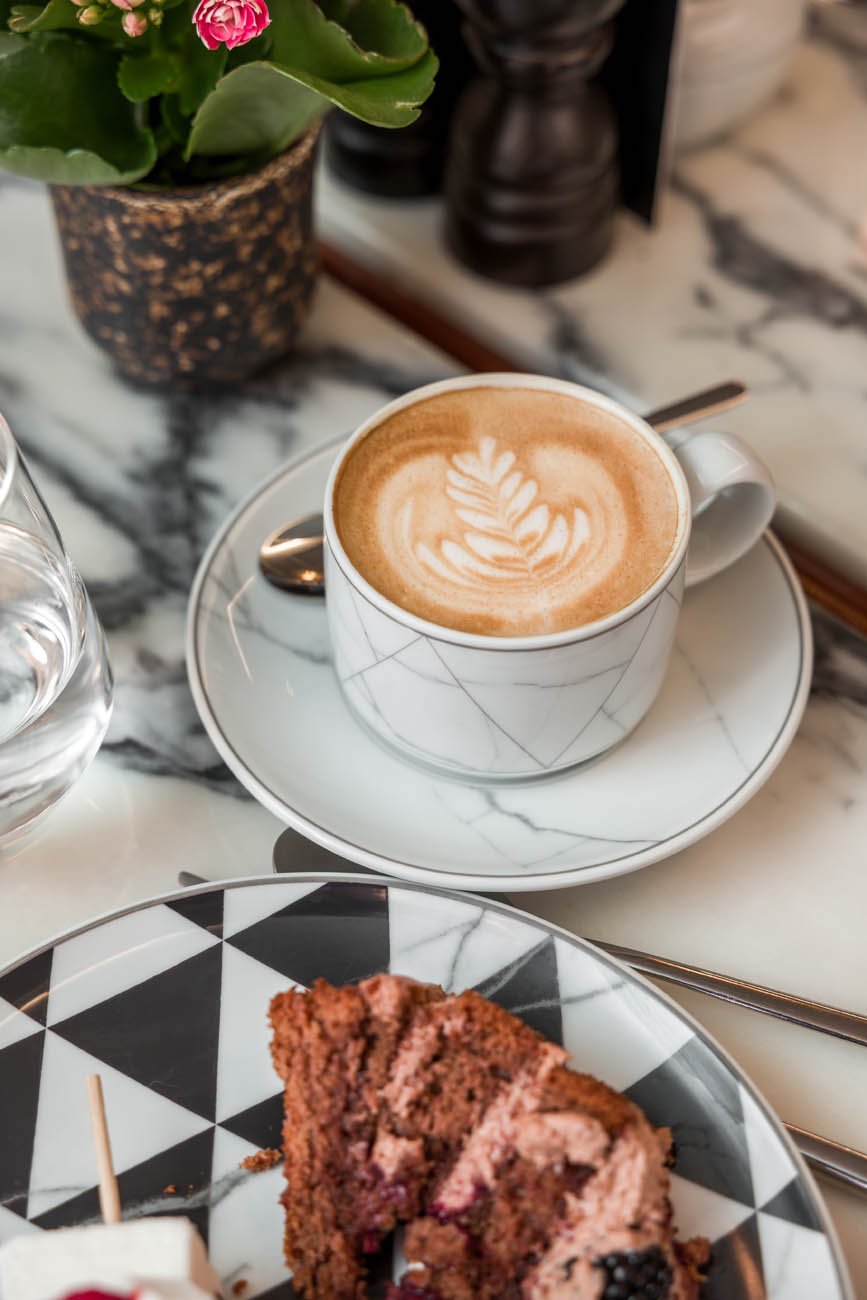 Cappuccino com arte de latte, acompanhado de uma fatia de bolo em prato decorativo preto e branco, em mesa de mármore, com planta de flores ao fundo.