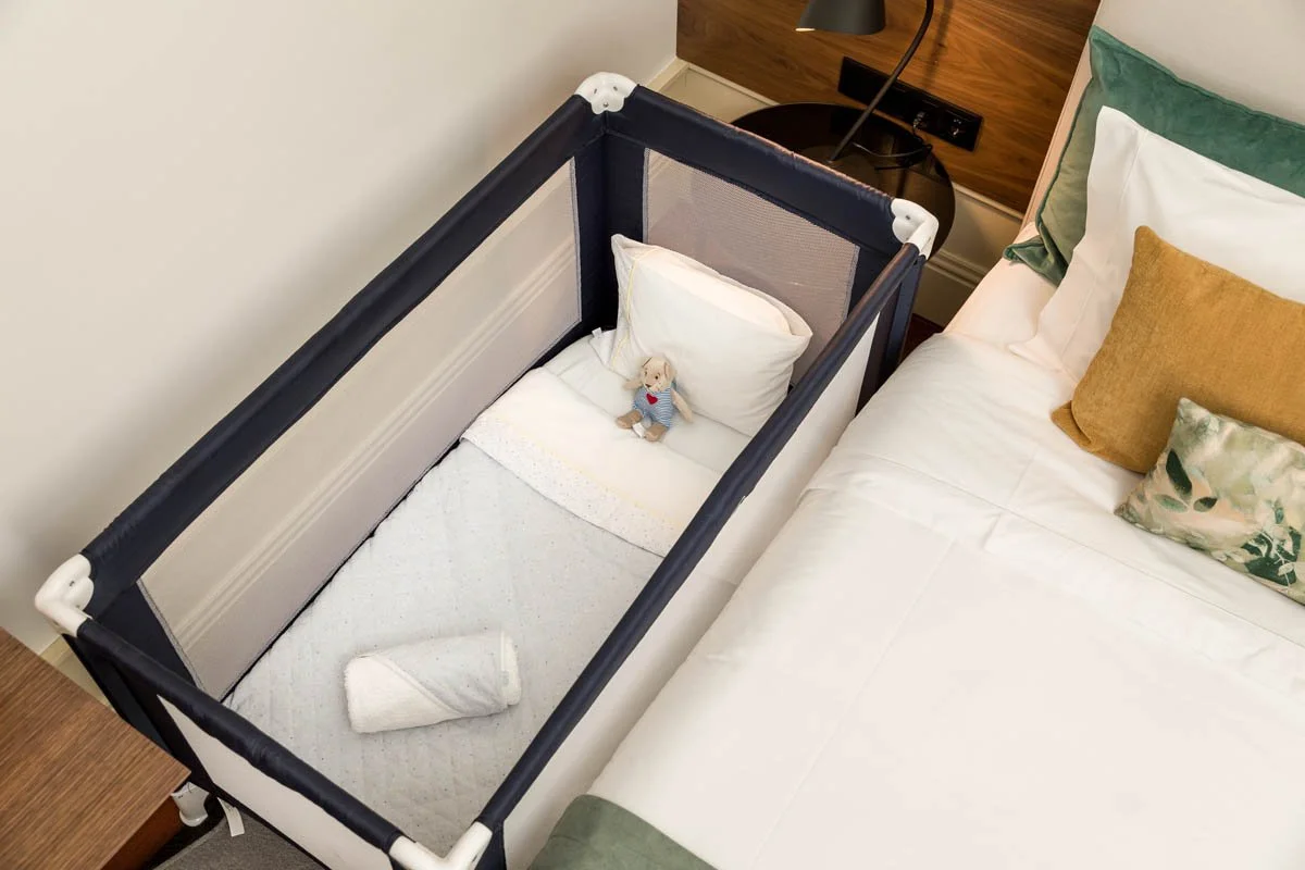 Berço ao lado de uma cama de casal com almofadas coloridas, na parede uma luminária e uma tomada