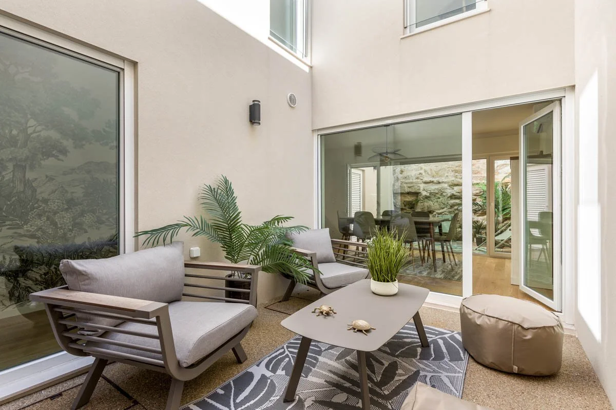 Varanda com duas cadeiras de descansoo, uma mesa com plantas, uma almofada e uma parede de vidro.