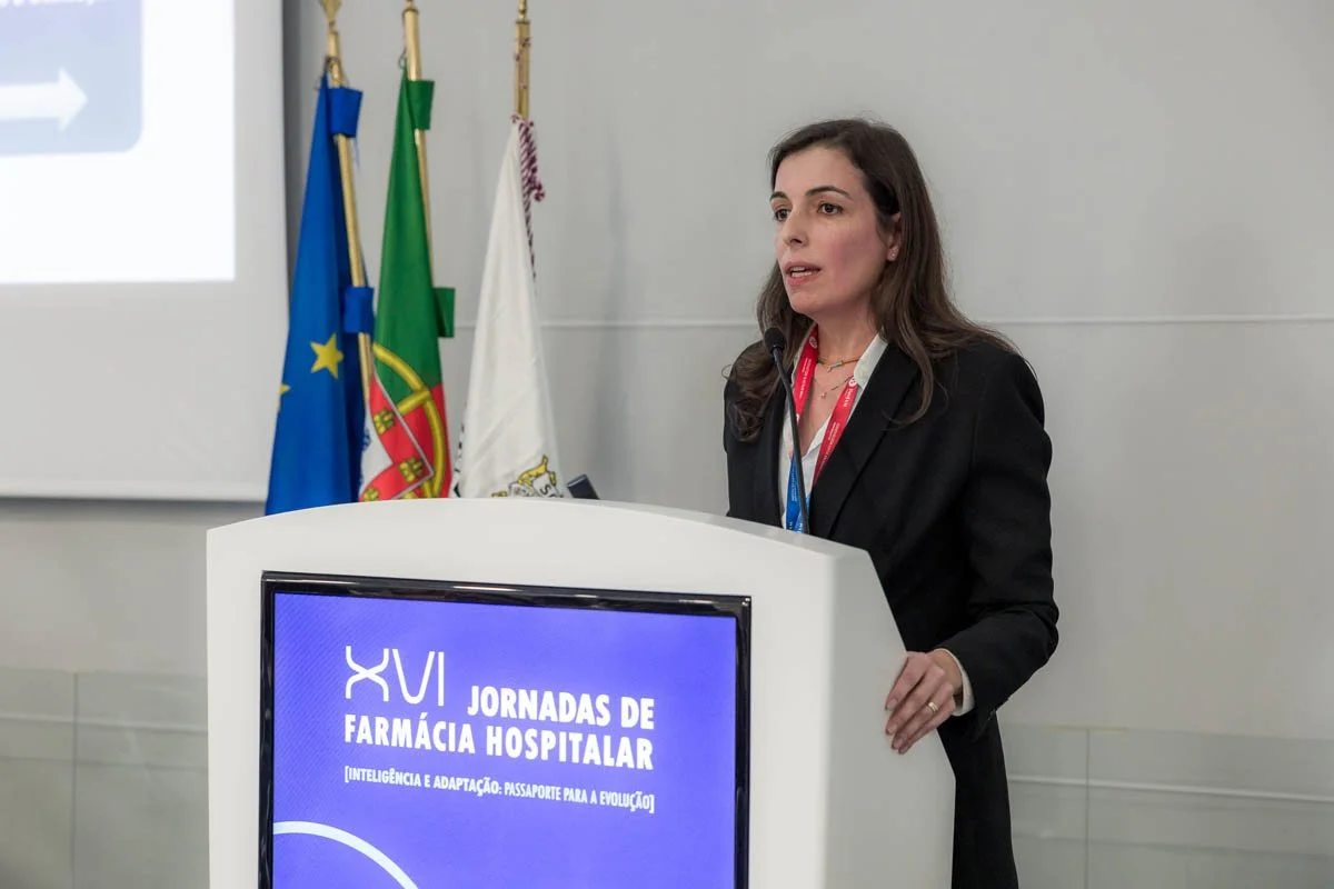 Mulher falando em um palco com uma tela ao fundo que diz "XVI Jornadas de Farmácia Hospitalar" em português, ao lado de bandeiras de Portugal e da União Europeia.