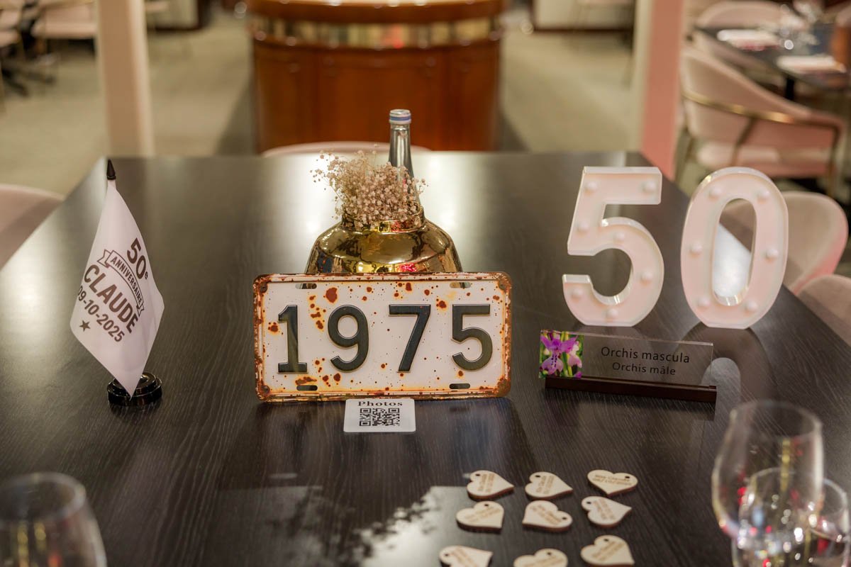 Decoração de mesa de aniversário de 50 anos, com um letreiro de números brancos de LED, uma placa com o ano 1975 e uma vela decorativa com flores e uma garrafa de vidro dourada ao fundo.