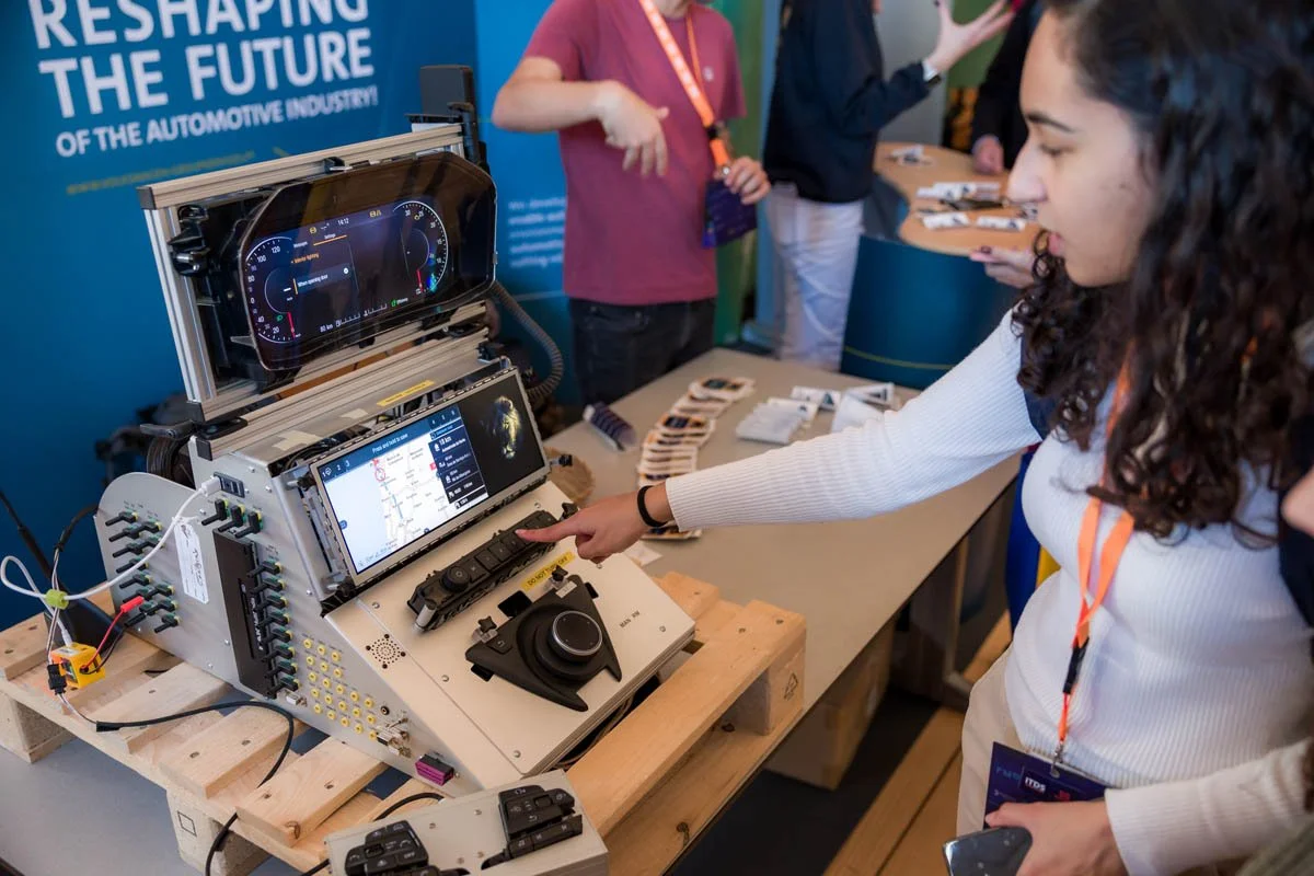 Pessoa interagindo com uma bancada de demonstração de tecnologia automotiva em uma feira, com um painel digital, controles e dispositivos eletrônicos, enquanto outras pessoas observam ao fundo.
