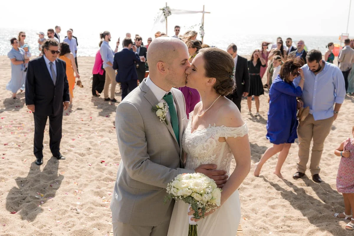 Casal de noivos beijando na areia durante casamento na praia, com convidados ao fundo e decoração com arco floral.