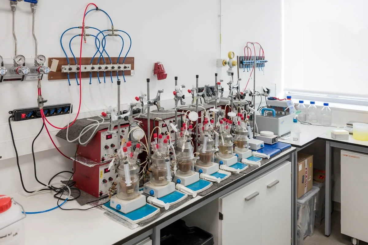 Laboratório de química com diversos bicos de gás conectados a frascos de vidro em uma bancada, equipamentos de laboratório e bottles de água ao lado.