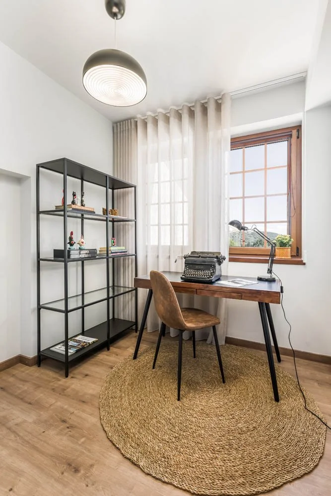 um escritório com uma mesa de madeira, uma cadeira de cor marrom escuro, uma prateleira de ferro preta com objetos decorativos, um tapete redondo de sisal, uma janela de rancho com cortinas brancas e uma luminária de teto em formato de meia-lua.