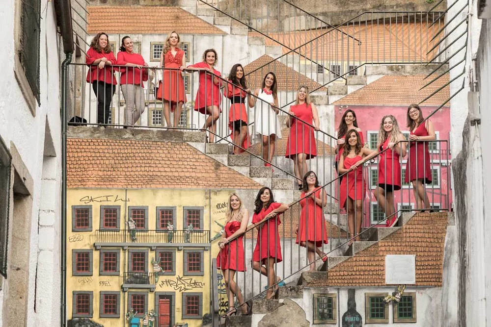 Grupo de mulheres em vestidos vermelhos e brancos, posando em escadaria de prédio com mural de casas coloridas ao fundo.