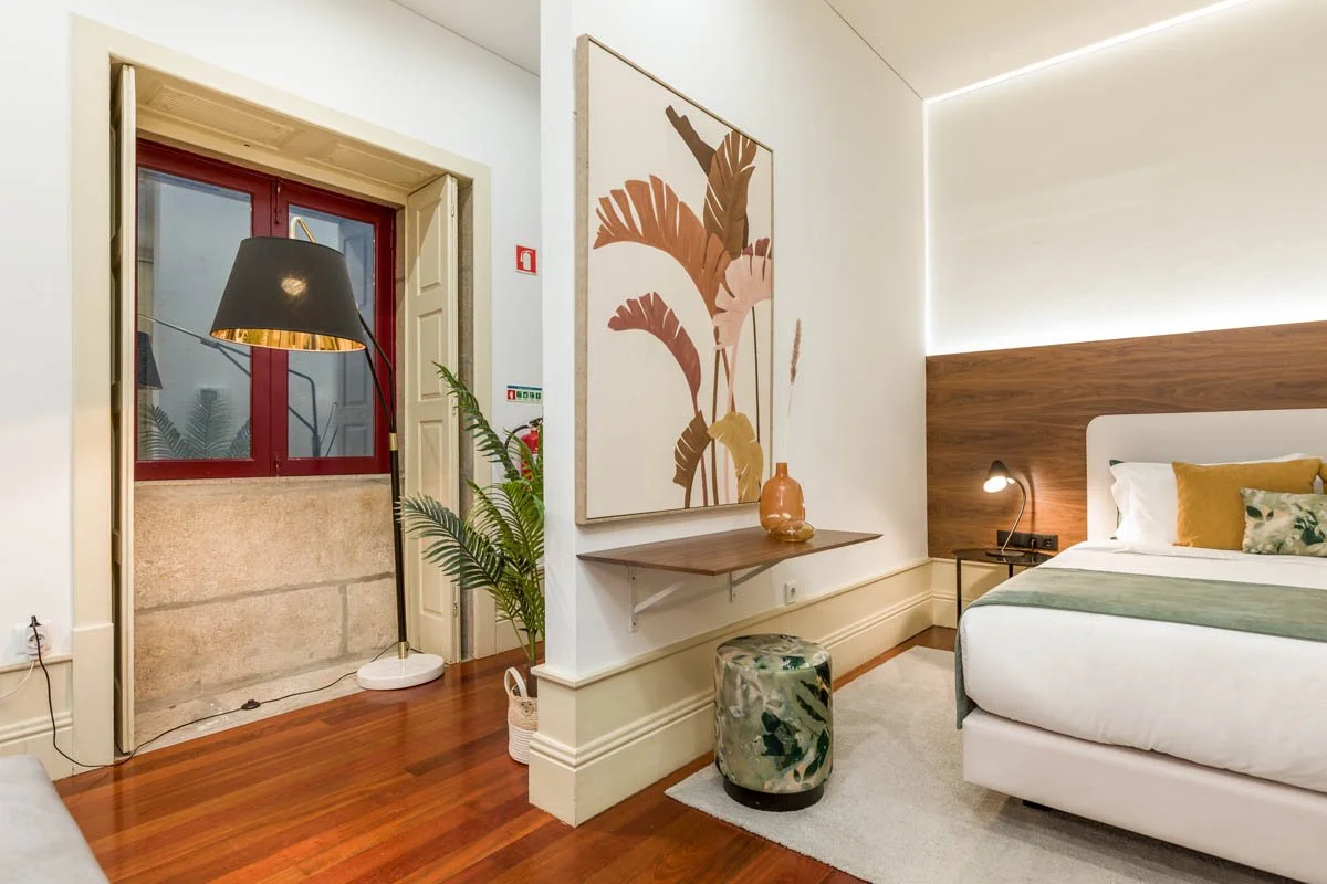 Quarto de hotel com cama de casal, cabeceira de madeira, abajur ao lado, parede com iluminação indireta, quadro com planta em tons bege e rosa, piso de madeira escura, janela com cortinas abertas, planta ao lado da janela e uma poltrona estampada na 