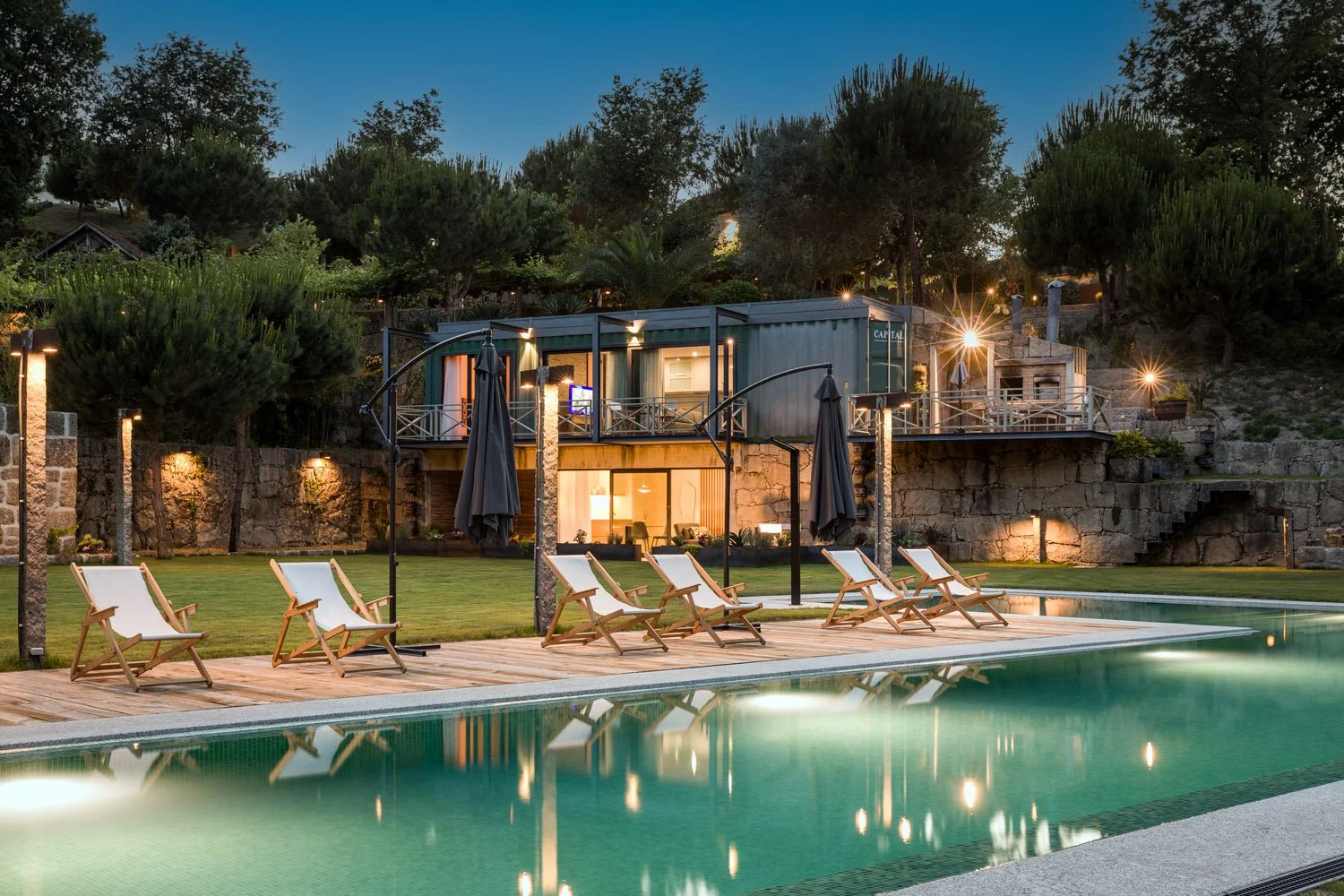 Cena de uma área de lazer ao entardecer com piscina, espreguiçadeiras de madeira com tecido branco, guarda-sóis pretos fechados, iluminação externa e uma casa moderna de dois andares ao fundo, cercada por árvores e vegetação.