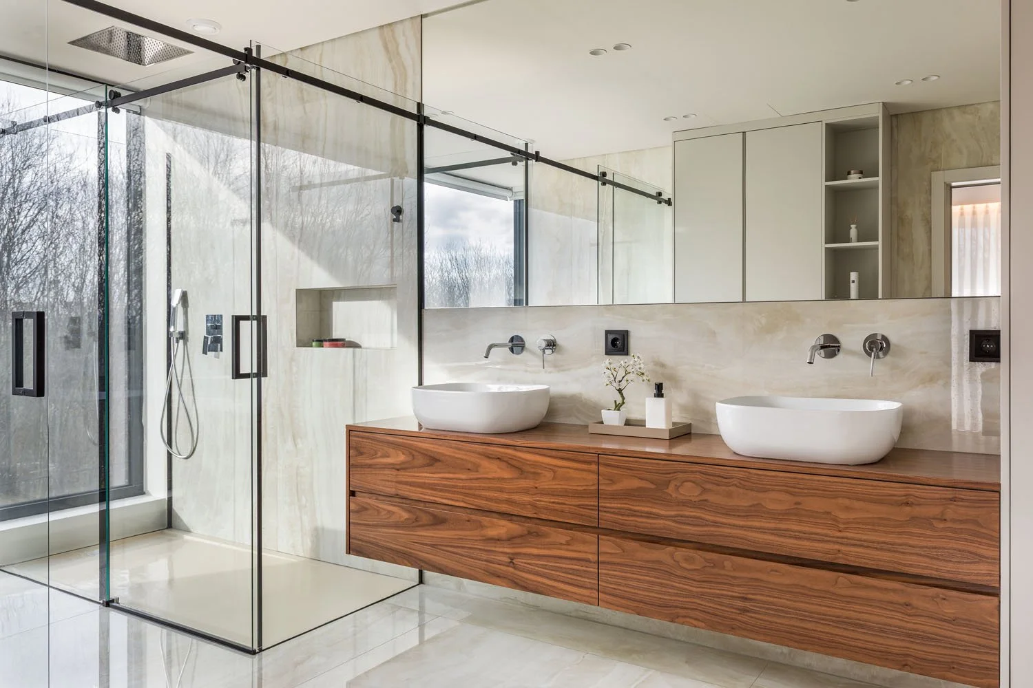 Banheiro com parede de mármore, cuba de vaso de cerâmica em uma bancada de madeira, espelho grande, espaço de ducha com vidro transparente, janela ao lado, decoração minimalista com uma planta e sabonete líquido.