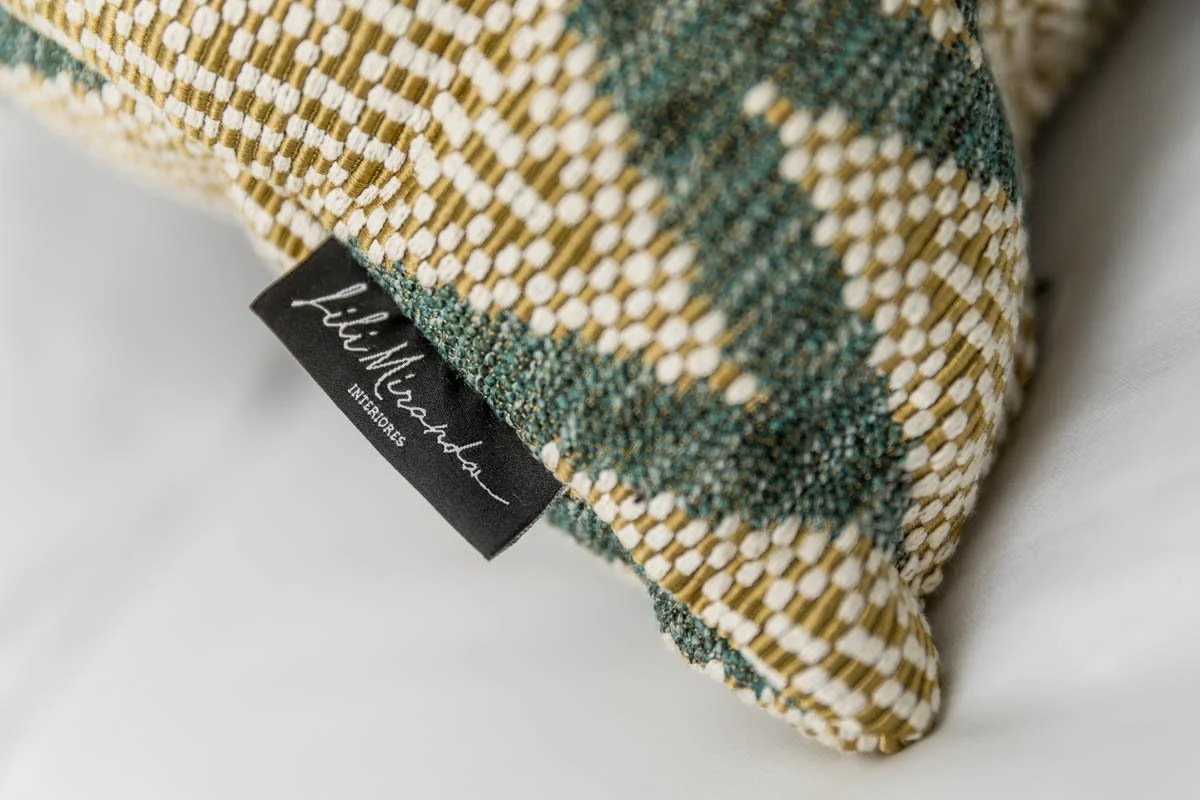 Detalhe de uma almofada com tecido de trama entrelaçada em cores bege, branco e verde, com etiqueta preta com escrita branca