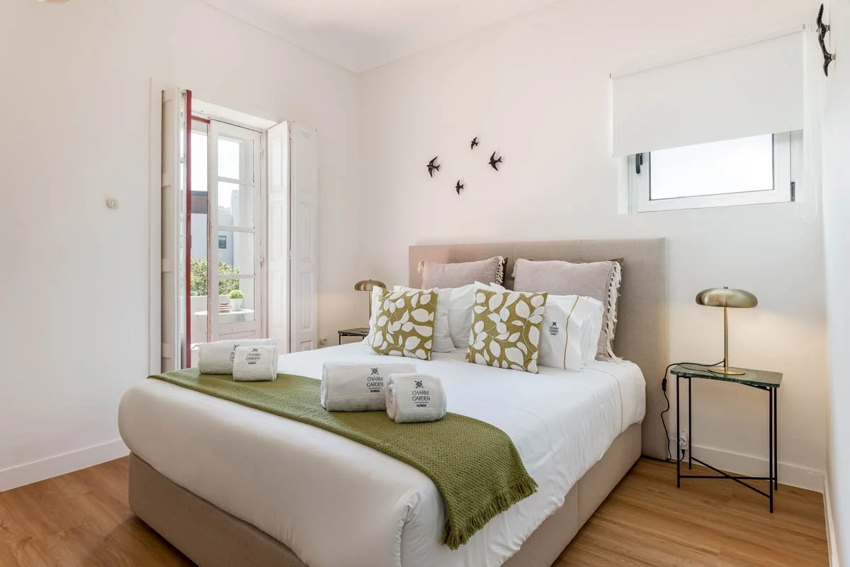 Quarto de hotel com cama de casal, travesseiros, cômoda ao lado, janelas abertas com cortinas brancas, decoração minimalista com pássaros na parede.