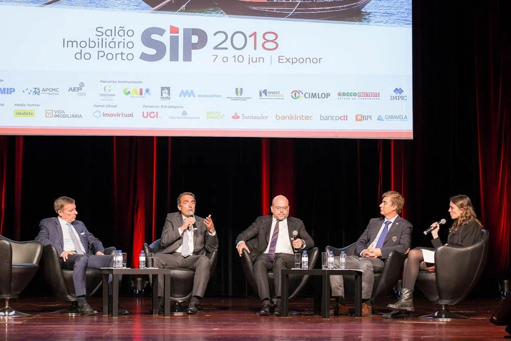 Painel de discussão com cinco pessoas sentadas em cadeiras de palco durante evento no Salão Imobiliário do Porto, com tela exibindo detalhes do evento atrás deles.
