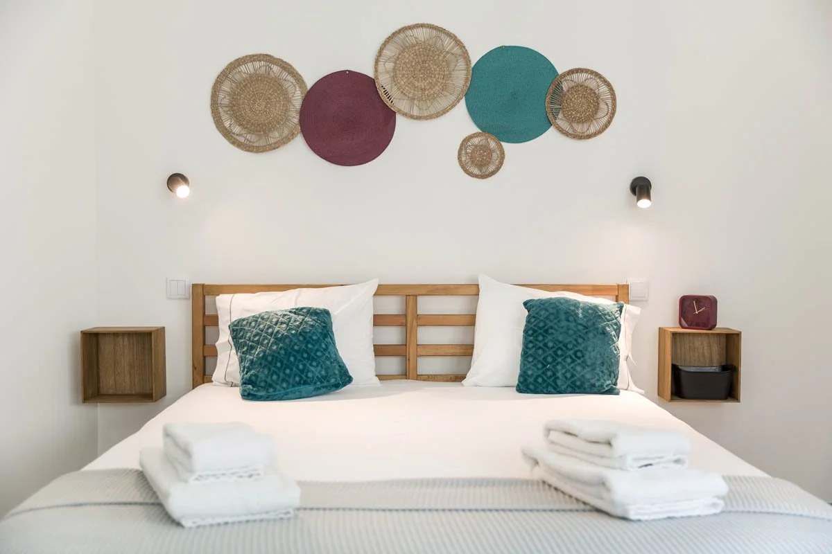 Cama de casal com cabeceira de madeira, duas almofadas verdes azuladas e dois conjuntos de toalhas brancas, decoração na parede com círculos de palha coloridos e dois focos de luz