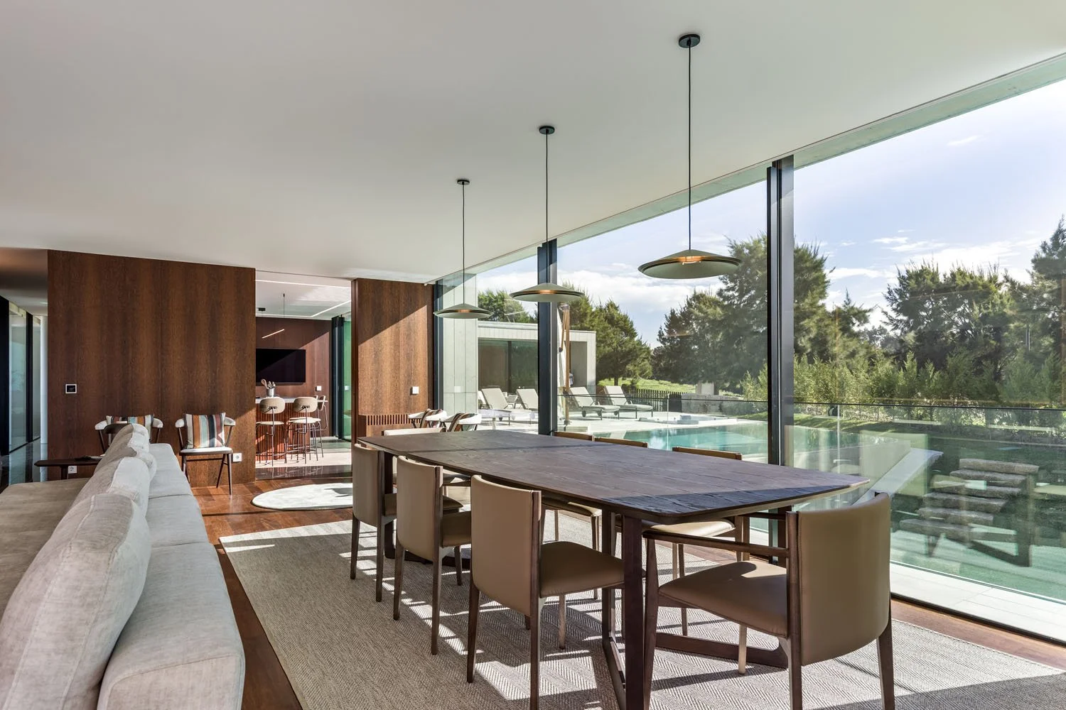 Sala de jantar moderna com mesa de madeira, cadeiras de couro bege, sofá branco, painel de madeira escura na parede, plantas ao ar livre, vista para piscina e área externa com espreguiçadeiras.