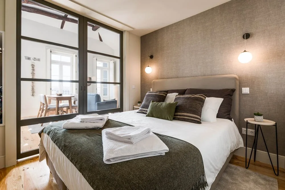 Quarto de hotel com cama de roupas brancas, almofadas decorativas e toalhas em cima. Há uma janela de vidro com porta de correr levando a uma varanda com mesa e cadeiras. O quarto tem parede acinzentada, iluminação com dois abajures ao lado da cama e