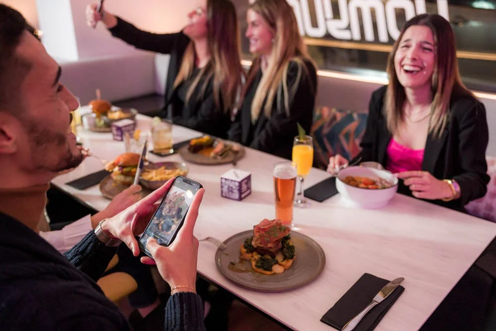 Grupo de cinco pessoas, quatro mulheres e um homem, sentados em uma mesa de restaurante, sorrindo e se divertindo enquanto uma mulher tira uma foto com o celular. A mesa está com pratos de comida, copos de bebida e utensílios.