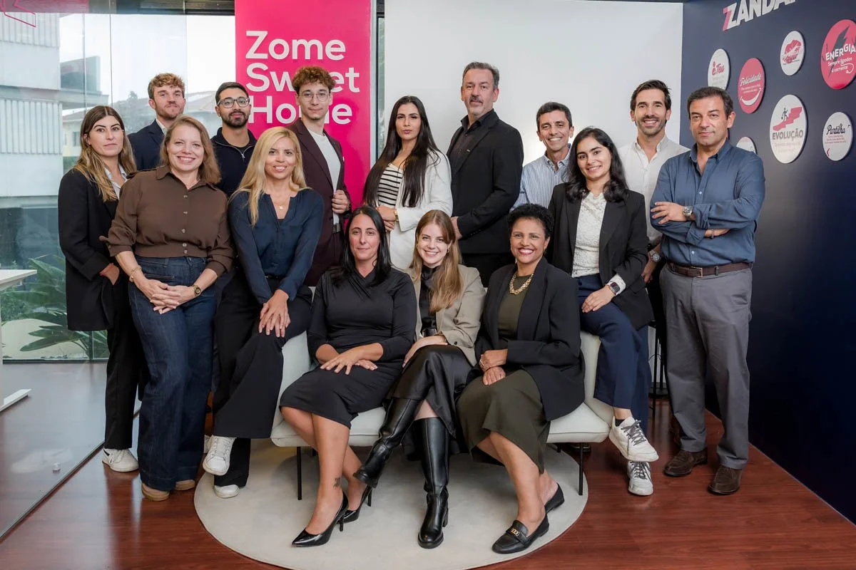 Grupo de pessoas posando para uma foto em um ambiente de escritório, com uma parede ao fundo contendo logotipos e um banner rosa com as palavras "Zome Smart Home".
