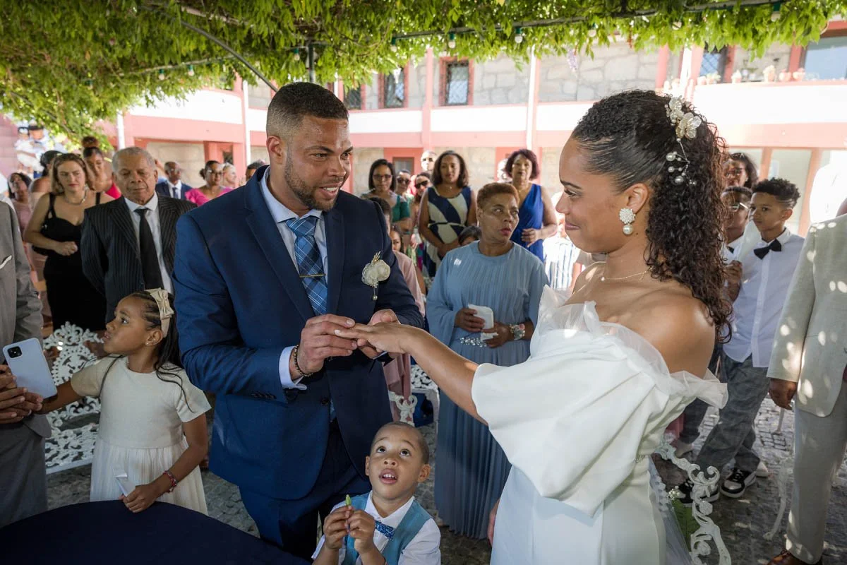 Casal trocando alianças durante uma cerimônia de casamento ao ar livre, rodeado por convidados.