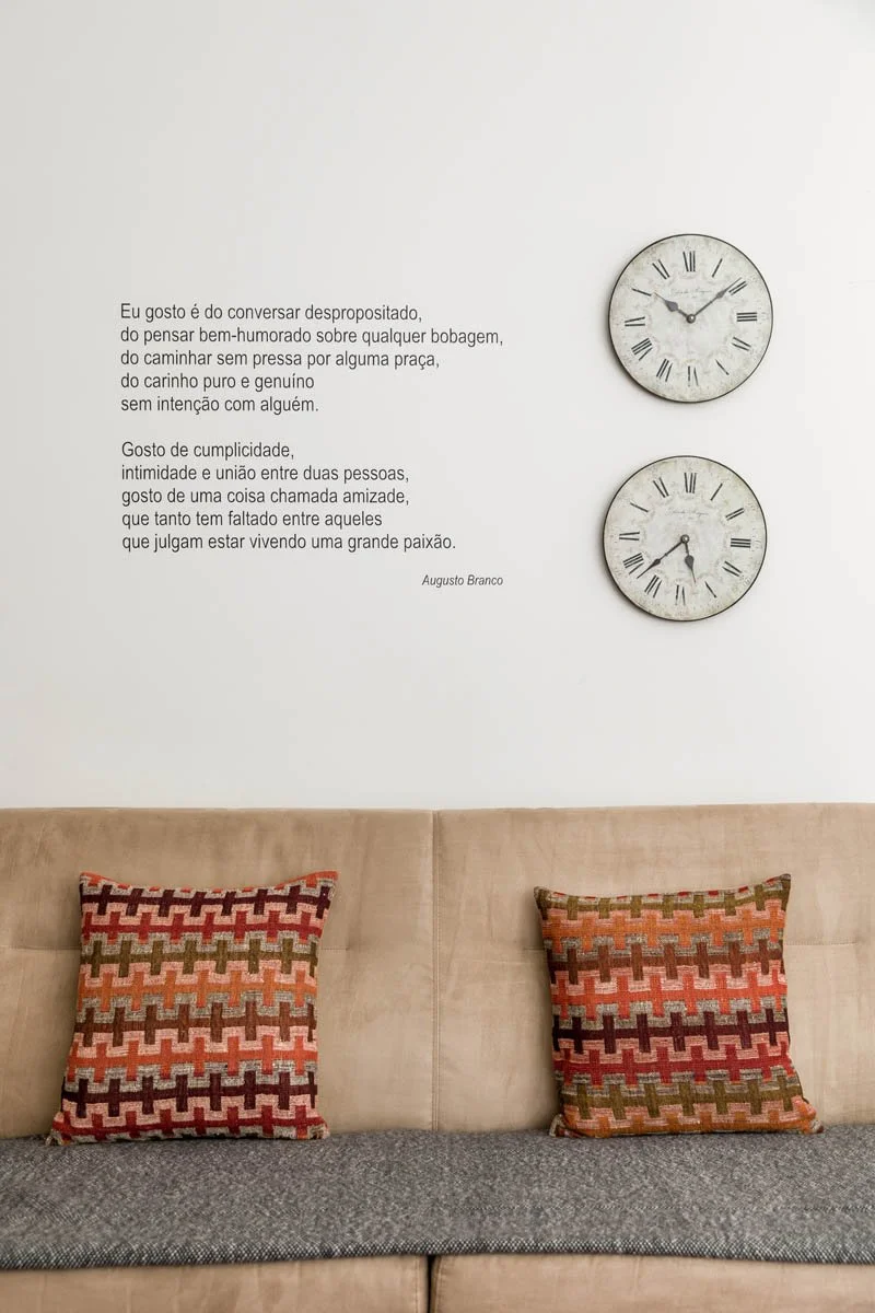 Sala com dois relógios de parede vintage, uma parede branca com uma citação de Augusto Branco, um sofá bege com dois almofadas decorativas de padrão geométrico em tons de vermelhos, verdes, rosa e preto.