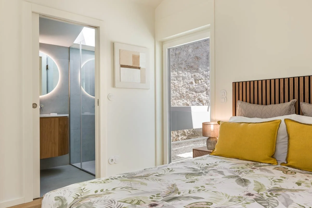 Quarto de cama com roupa de cama com estampa floral, travesseiros cinza e amarelos, parede branca, janela com vista para uma parede de pedra e banheiro com iluminação de LED ao redor do espelho.