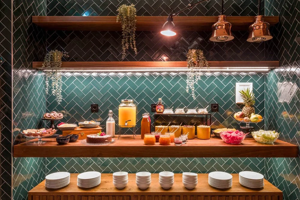 Tábua de café da manhã com frutas, sucos, iogurte e doces em uma parede de azulejos verdes.