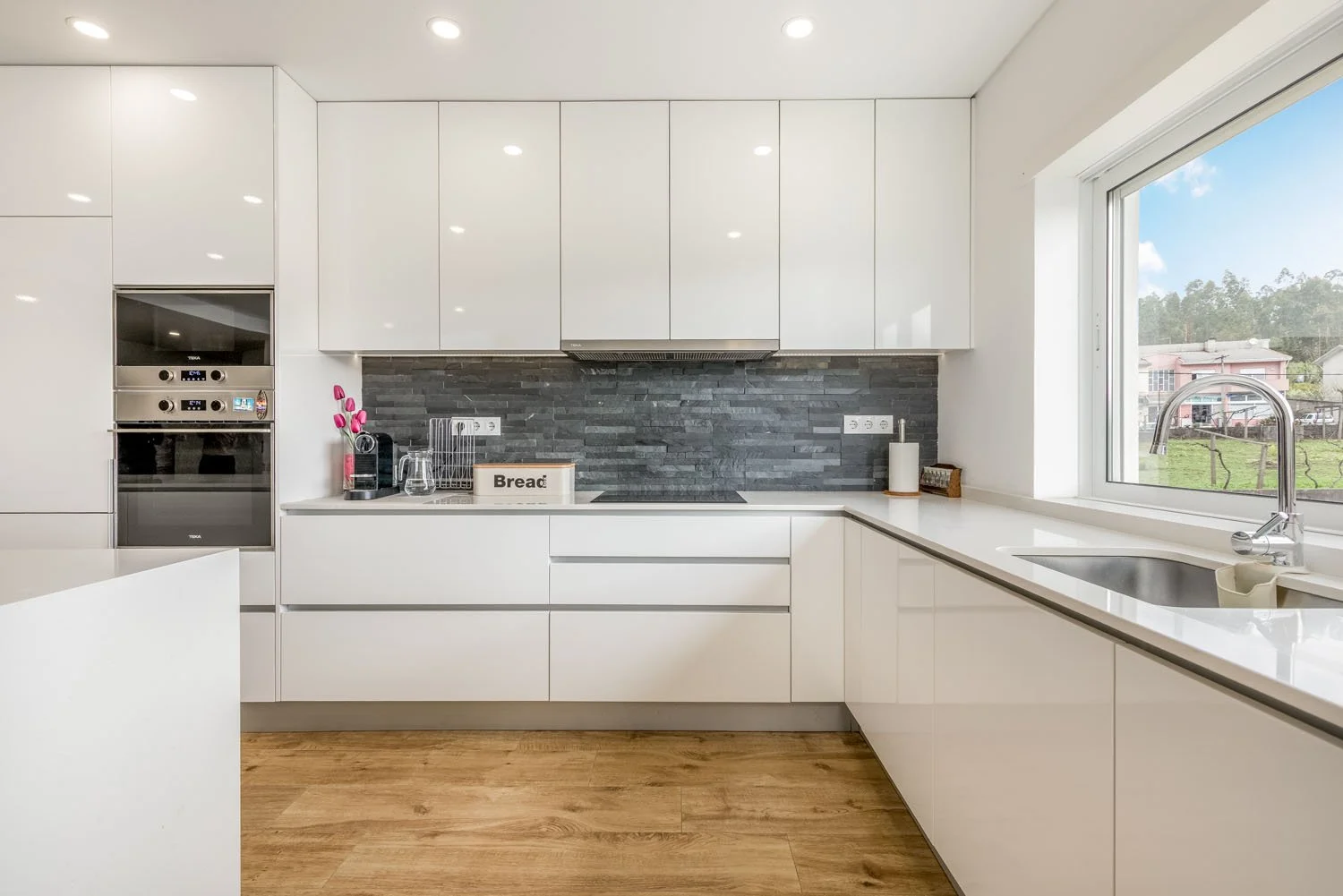 Cozinha moderna com armários brancos, bancada de mármore branco, parede de revestimento escuro, janela grande, pia com torneira, piso de madeira clara, eletrodomésticos embutidos e decoração minimalista.