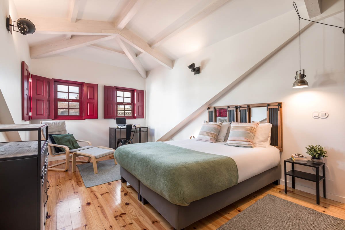 Quarto com cama, cabeceira de madeira escura, roupa de cama branca, cobertor verde, dois travesseiros listrados, parede branca, janelas com persianas vermelhas abertas, móveis de madeira e preto, cadeira de escritório, escrivaninha, poltrona de fibra