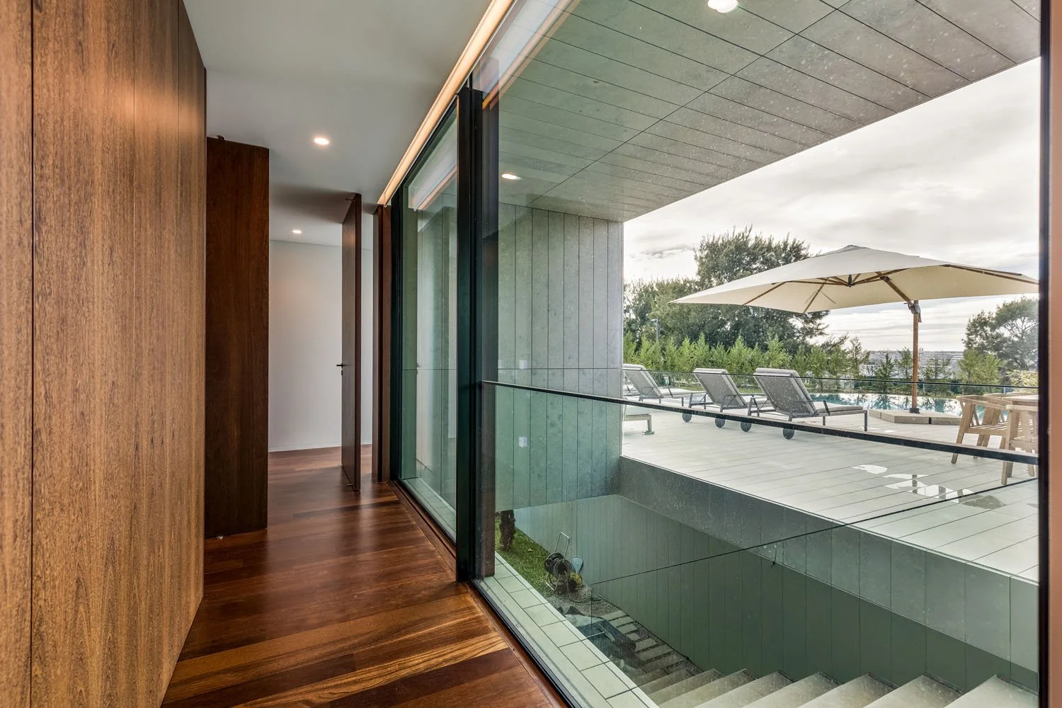 Interiores de uma casa com piso de madeira, parede de madeira escura e uma grande janela de vidro que dá para uma varanda com espreguiçadeiras, guarda-sol e uma mesa de jardim, ao ar livre.