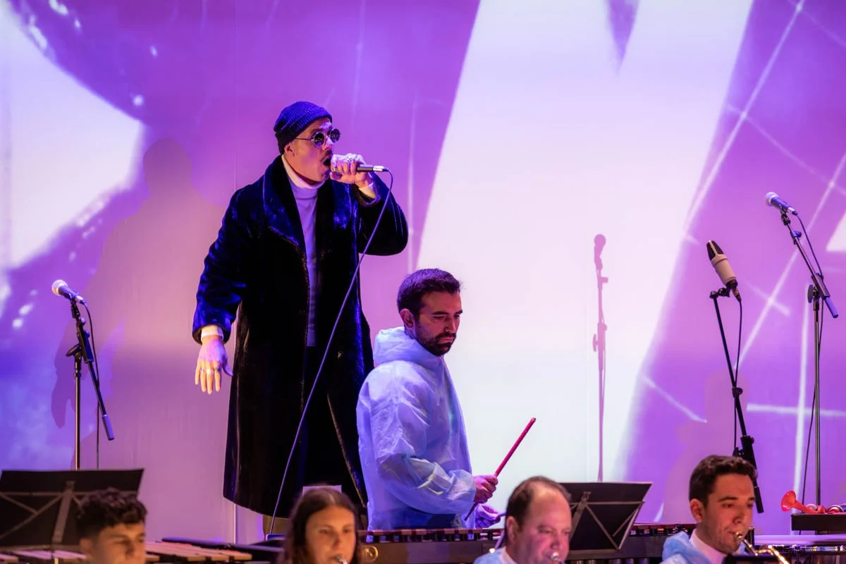 Performers on stage, one singing into a microphone, others playing instruments, with a colorful abstract background.