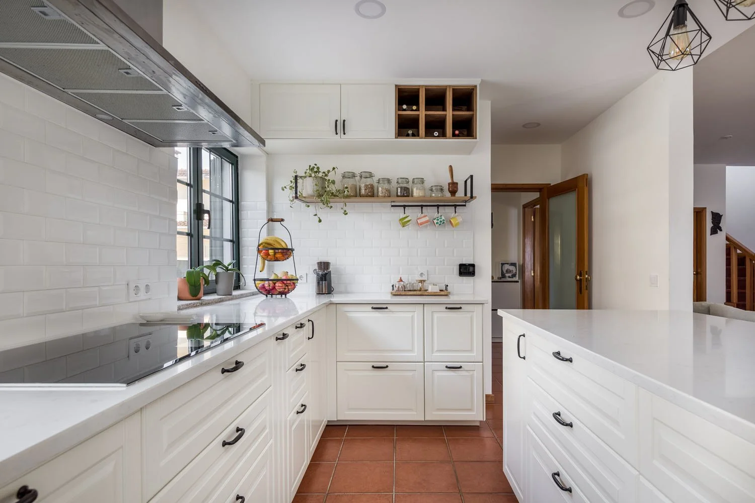 Cozinha moderna com armários brancos, bancada de mármore, janelas ao lado da pia, decoração com plantas, potes de vidro e utensílios na parede, piso de piso de tijolinho.