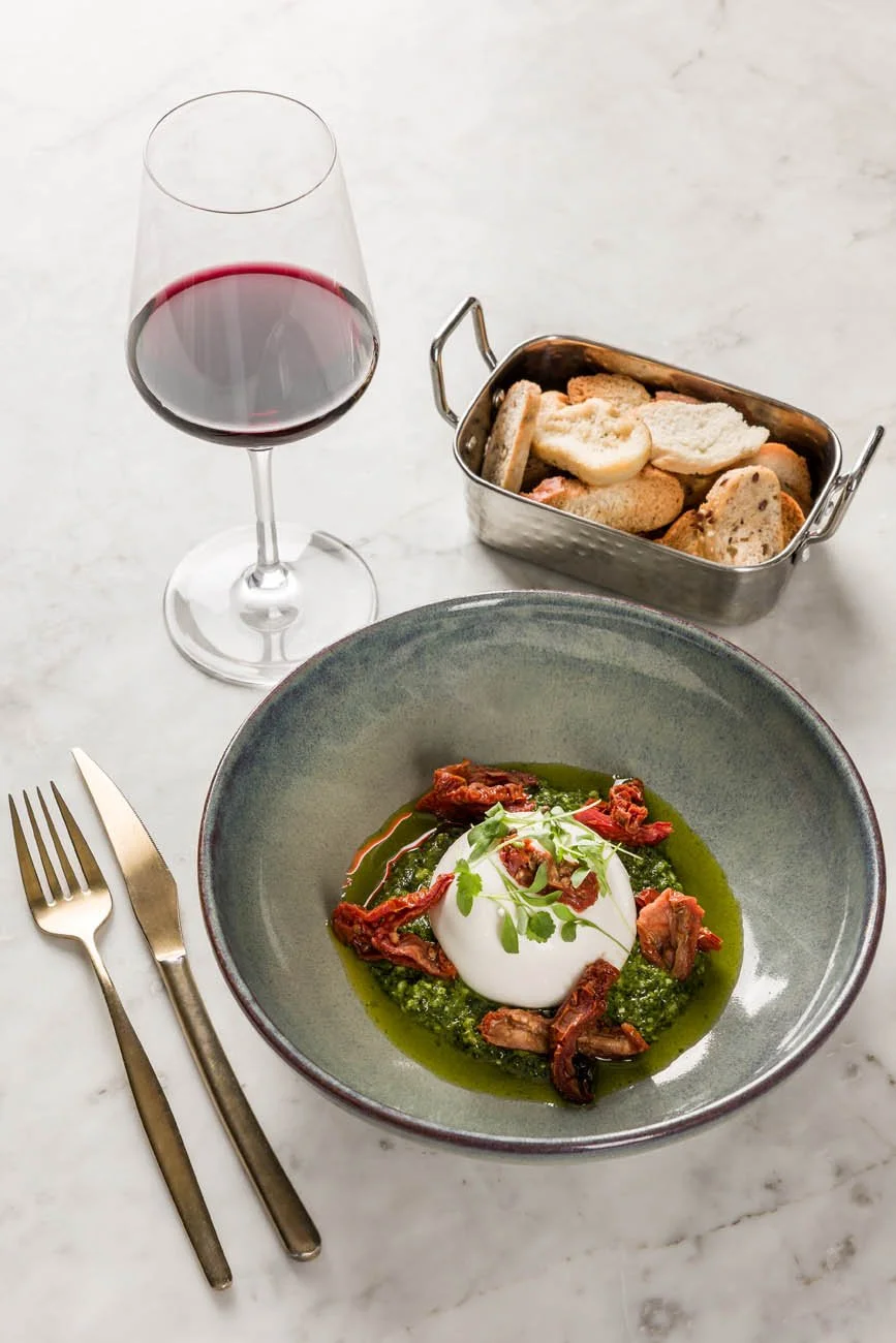 Prato com ovo pochê, molho verde, carne desfiada e ervas, ao lado de uma taça de vinho tinto, pão e queijo em uma bandeja.