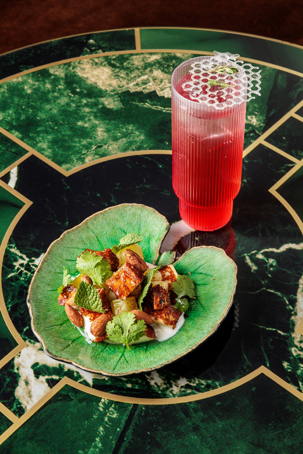 Prato com pedaços de carne, folhas de hortelã e coentro, servido em uma tigela de cerâmica verde, ao lado de um copo de drink rosa com decoração de melancia e hortelã, sobre mesa de mármore verde escuro com detalhes dourados.