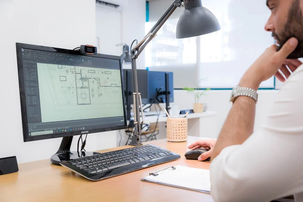Pessoa trabalhando em frente a um monitor de computador com um desenho de planta baixa. Está em um escritório, com uma luminária, uma caneca, um notebook e uma planta ao fundo.