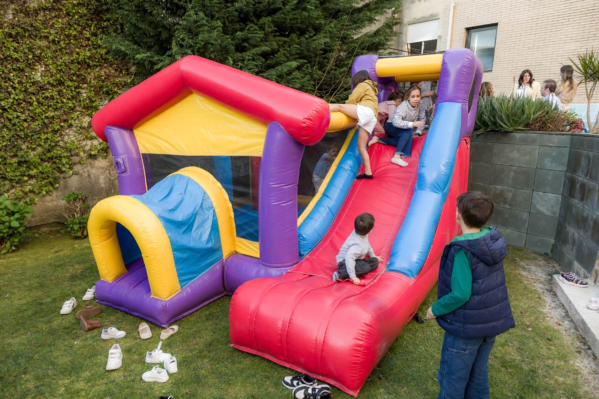 Crianças brincando em escorregador inflável colorido no quintal, com adultos observando.