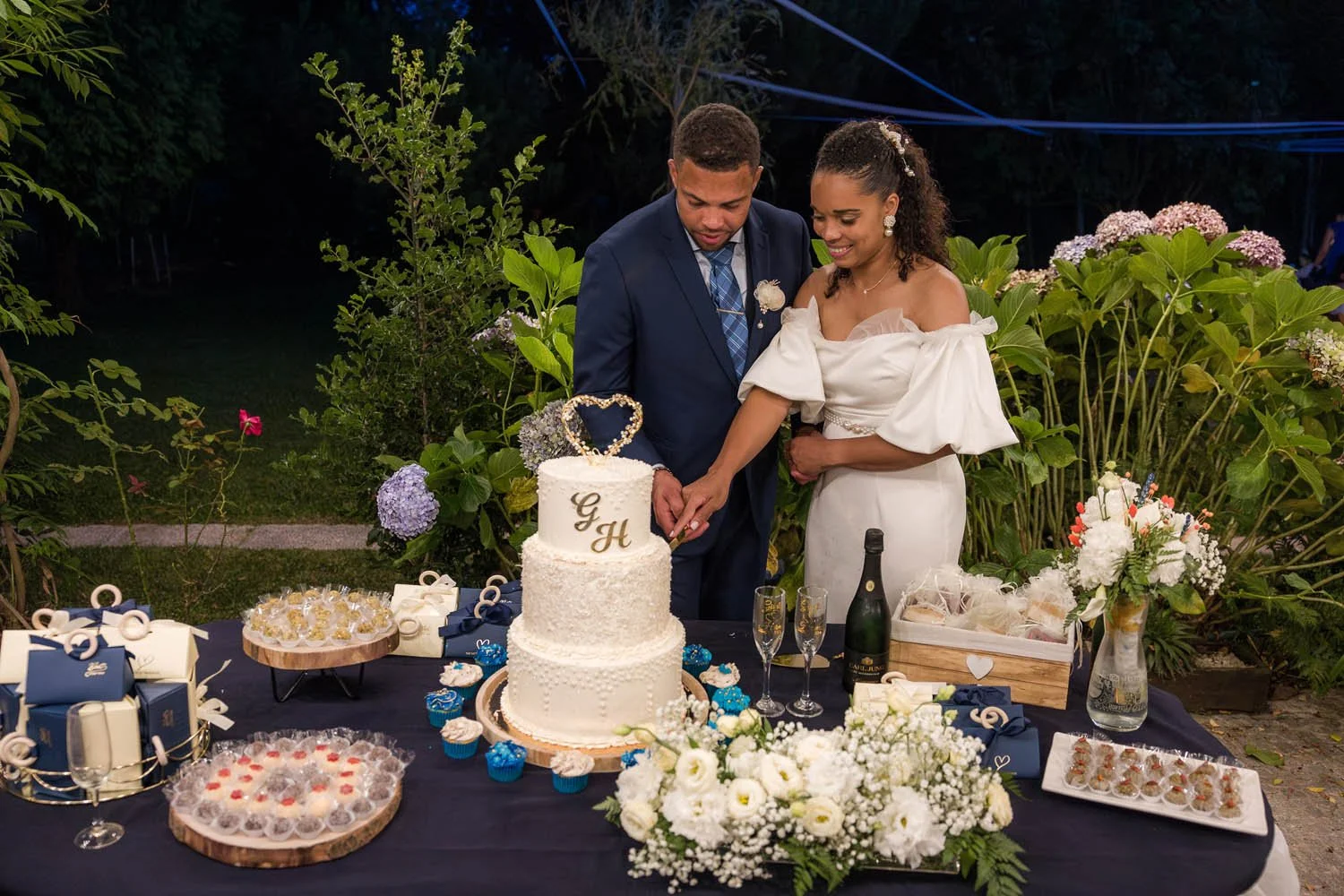 Casal de noivos cortando o bolo de casamento ao pôr do sol com decoração de flores brancas e doces na mesa, ao ar livre.