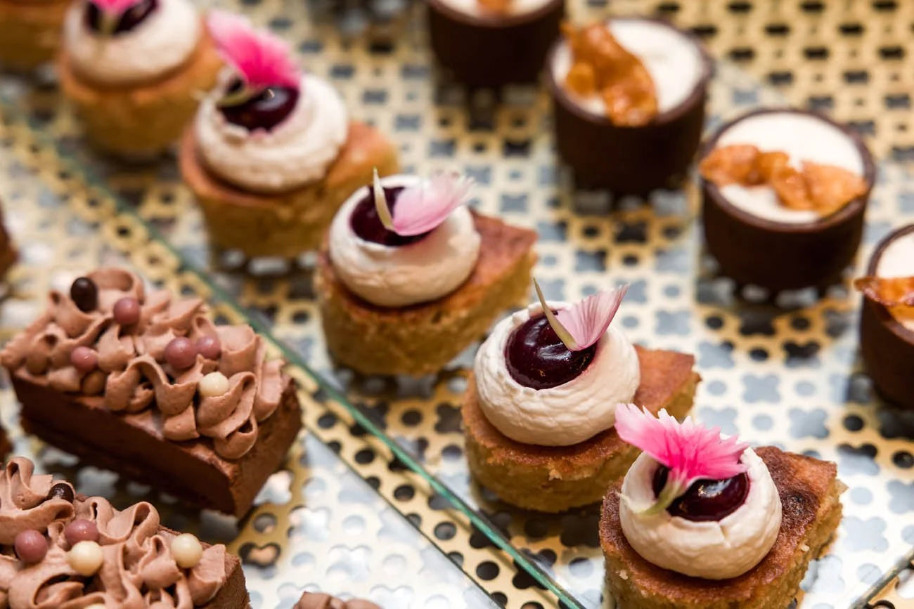 Diversos doces finos decorados com creme, frutas, flores comestíveis e confeitos em uma bandeja de metal.