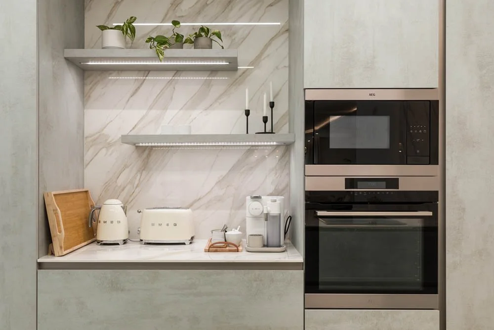 Cozinha moderna com eletrodomésticos brancos, micro-ondas e forno embutidos, prateleiras com plantas e vela, bancada limpa, parede com mármore claro.