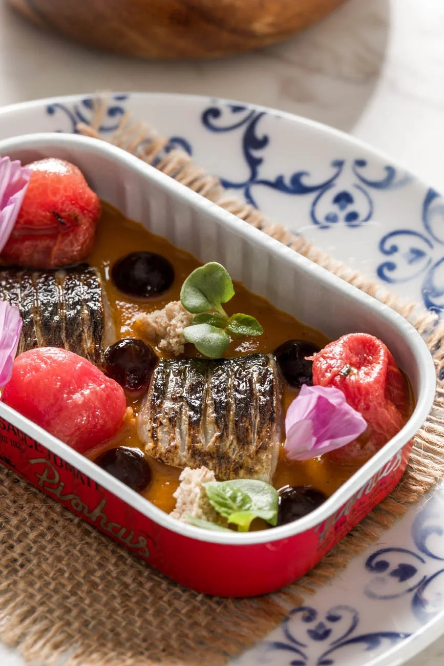 Prato de peixe com molho dourado, acompanhado de frutas vermelhas, flores comestíveis e folhas verdes, servido em uma travessa decorada.