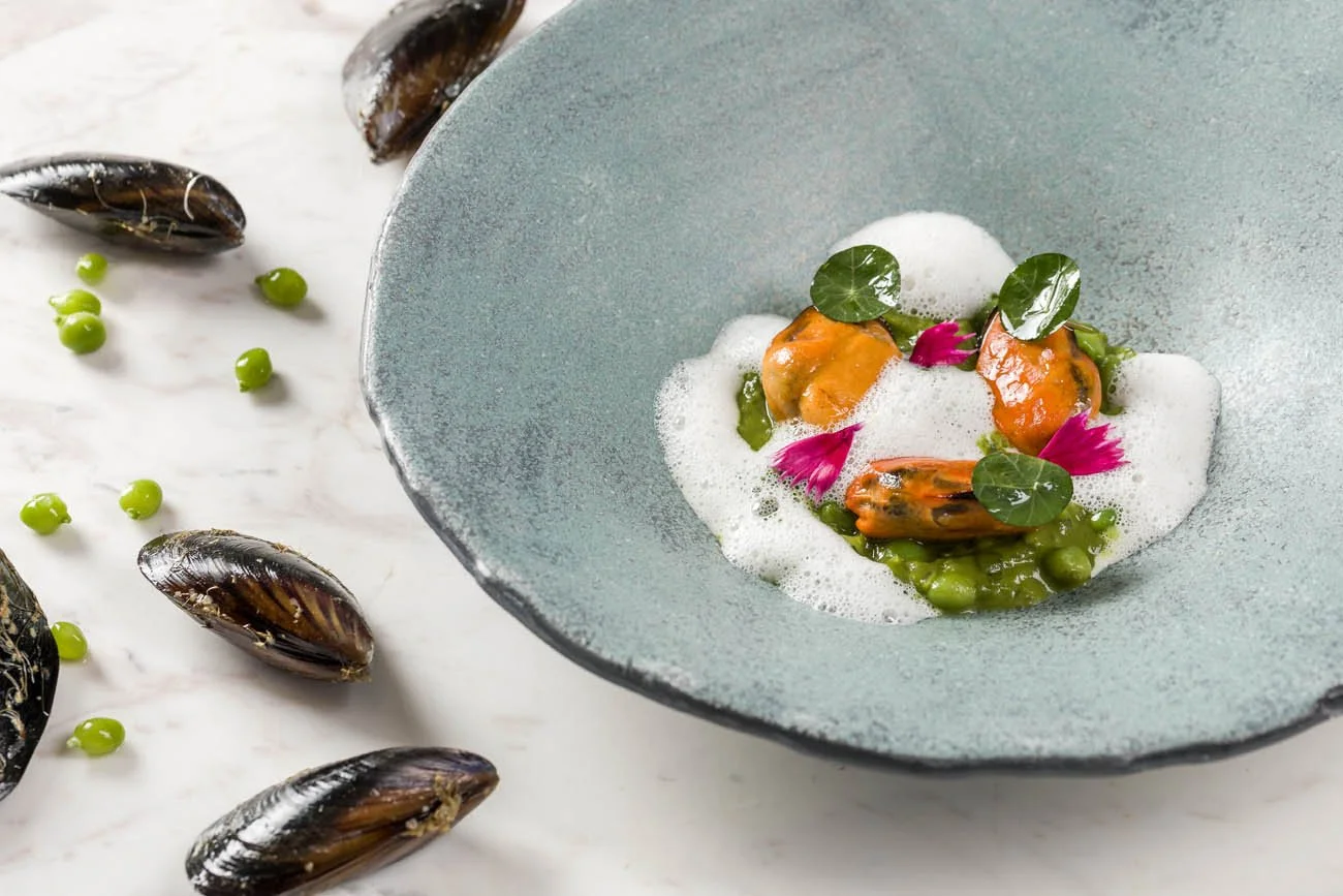 Prato de culinária sofisticada com moluscos, ervilhas, folhas verdes, flores roxas, espuma e um prato azul com fundo de superfície branca com mexilhões e ervilhas espalhadas.